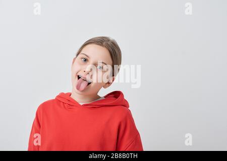 Drôle de jeune fille dans le sweat-shirt rouge montrant la langue tout en grimaçant devant l'appareil photo contre le fond blanc isolément Banque D'Images