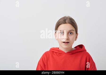 Une adolescente effrayée ou surprise dans un sweat à capuche rouge se tenant devant l'appareil photo contre un fond blanc et vous regardant Banque D'Images