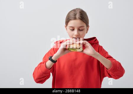 Jeune fille dans le hoodie rouge tenant appétissant hamburger par sa bouche ouverte tout en allant le manger contre fond blanc dans l'isolement Banque D'Images