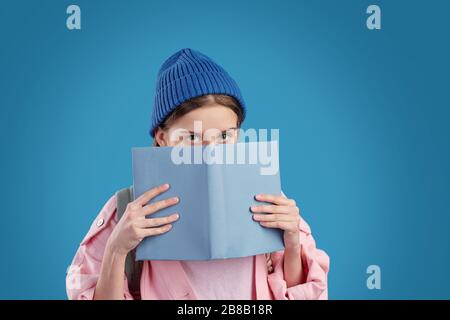 Une adolescente timide dans un bonnet bleu et une veste en denim rose qui se déchit du livre ouvert près du visage tout en se tenant devant l'appareil photo Banque D'Images