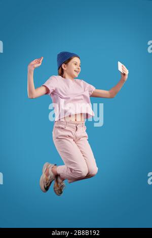 Une adolescente ecstatic en tenue de nuit tenant son smartphone devant elle-même tout en faisant un selfie en sautant sur le fond bleu Banque D'Images