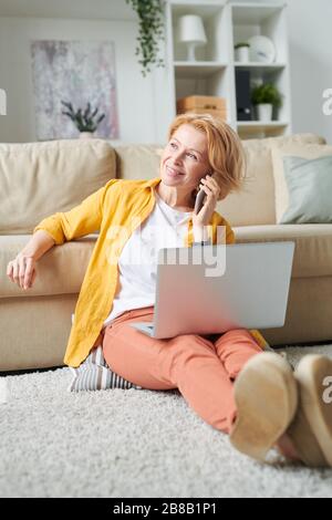 Femme d'affaires blonde et gaie avec PC portable et smartphone consultant clients tout en travaillant à distance à la maison Banque D'Images