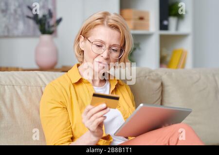 Femme blonde mûre dans les lunettes et vêtements de casual regardant la carte de crédit tout en allant faire des achats en ligne pendant la quarantaine à la maison Banque D'Images