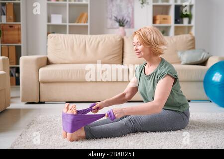 Heureuse femme mûre dans les leggins grises et t-shirt assis sur le plancher du salon et faire de l'exercice pour les jambes pendant la quarantaine à la maison Banque D'Images