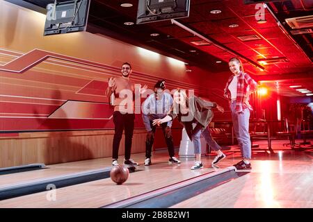 Groupe de jeunes amis joyeux de diverses ethnies debout par bowling tout en obne d'eux lancer la balle pendant le jeu Banque D'Images
