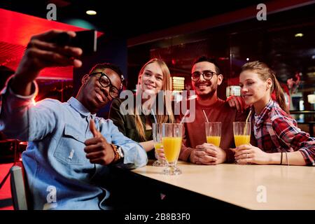 Jeune homme africain avec smartphone montrant le pouce tout en faisant selfie avec ses amis par table dans un café au centre de loisirs Banque D'Images