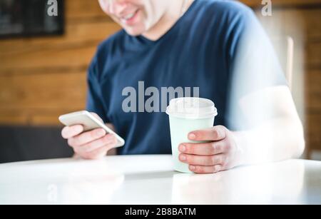 Un homme heureux utilisant un smartphone dans un café ou à la maison. Un gars souriant tenant un téléphone portable et une tasse à café en papier. SMS, achats en ligne ou navigation sur Internet. Banque D'Images