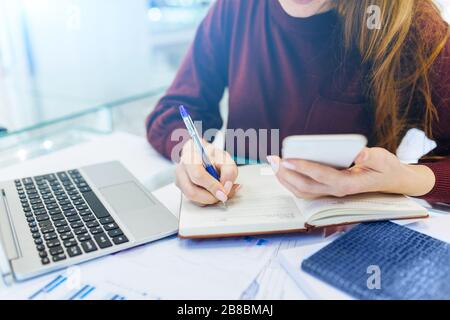 Une femme d'affaires travaille à la maison, au travail à distance à la maison, avec un ordinateur portable et un ordinateur portable, prend des notes sur le téléphone. Banque D'Images