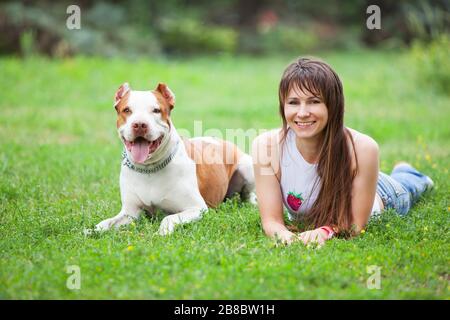 Vue de face d'une belle femme souriante reposant avec un adorable taureau de fosse sur l'herbe dans le parc. Charmante fille passe du temps avec quatre pattes frit en plein air. Con Banque D'Images