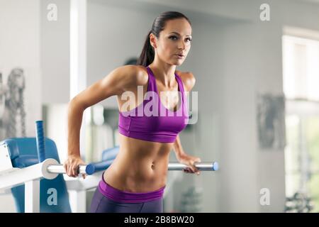 Vue latérale de la belle dame sportive dans les vêtements de sport utilisant la tour de puissance dans la salle de sport moderne. Charmante fille avec un corps bien en forme faisant de l'exercice de pull-up. Conceps Banque D'Images