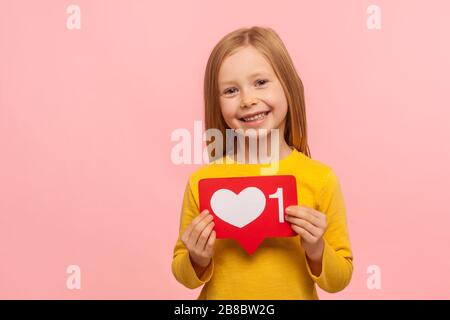 Contenu enfant populaire, blogging. Portrait de charmante jeune fille d'âge préscolaire tenant le coeur comme l'icône des médias sociaux et regardant l'appareil photo avec le sourire toothy. Banque D'Images