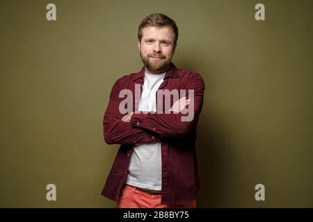 Calme, sympathique homme caucasien avec une barbe est barrée les bras croisés, souriant mignon et regardant l'appareil photo, sur un fond vert. Banque D'Images