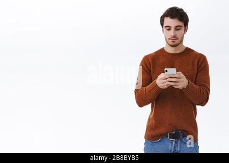 Jeune homme calme et détendu au beau look avec barbe, utilisant le téléphone mobile sans visage de poker d'émotions, SMS, appeler un ami ou envoyer des messages, utiliser Banque D'Images