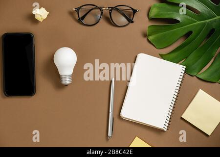 Idées inspiration concepts avec ampoule, stylo, smartphone moderne, feuilles de monstera, lunettes, ordinateur portable avec espace de copie sur fond brun. Modèle Banque D'Images