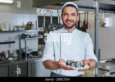 Sushis au restaurant. Il se tient dans la cuisine commerciale et tient son plat tout en regardant l'appareil photo avec un sourire. Vue de face Banque D'Images