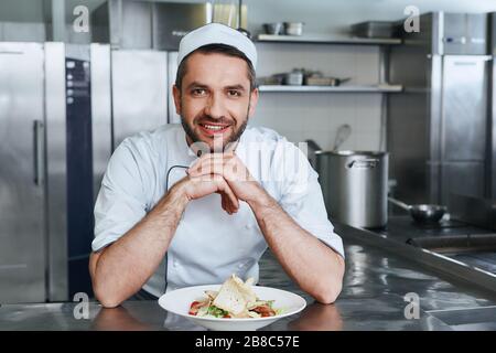 Un cuisinier souriant se tenant au-dessus du plat et regardant l'appareil photo. Chef caucasien préparant un délicieux repas dans la cuisine du restaurant Banque D'Images