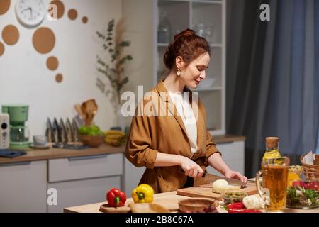Jeune femme coupant du fromage sur la planche à découper à la table, elle cuisine dans la cuisine domestique Banque D'Images