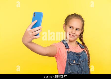 Portrait de charmante petite fille avec tresse dans des salopettes en denim souriant et prenant selfie sur le téléphone mobile, communiquer avec les parents sur appel vidéo. ind Banque D'Images