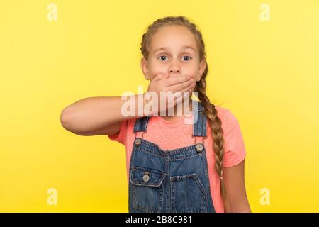 Je ne le dirai pas ! Portrait de la petite fille effrayée avec tresse dans des salopettes en denim couvrant la bouche avec la main, peur de dire secret, enfant terrifié de parler. Dans Banque D'Images