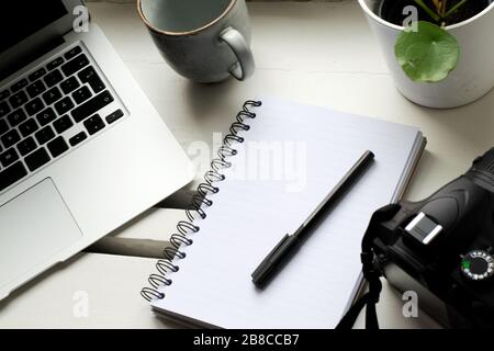 Travailler de la maison avec un lieu de travail à côté de la fenêtre avec ordinateur portable, appareil photo, usine, téléphone mobile, tasse de café, ordinateur portable et stylo sur la table Banque D'Images