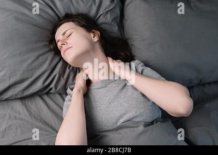 Une jeune femme malheureuse et fatiguée qui se couche au lit le matin ressent une douleur dans le cou après le sommeil nocturne, fait un massage des muscles tendus du cou, exsuce la souffrance des filles Banque D'Images