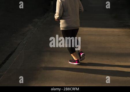 Vue arrière d'une femme en surpoids qui s'exécute dans un cadre lumineux sombre l'après-midi dans un cadre urbain et asphalté à la périphérie d'une rue de banlieue de la ville Banque D'Images