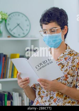 Une femme faisant des recherches sur Covid-19, une jeune femme lit un livre tout en portant un masque pour éviter la contagion, en arrière-plan une bibliothèque avec une horloge Banque D'Images