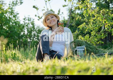 Vacances d'été dans le jardin, femme jardinier assis sur de l'herbe fraîchement coupée, se reposant femme avec des boissons fraîches aux herbes naturelles de baies Banque D'Images