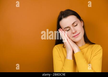 Jeune femme gaie avec le visage serein heureux tend à dormir, se penche sur les mains, a le sourire tendre, prend la sieste, porte le chandail jaune Banque D'Images