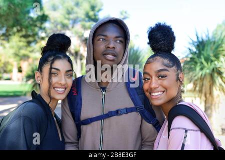 Un homme d'Afrique américain d'âge universitaire dans le hoodie entre deux coeds de couleur féminine avec des cool-dos à la mode sur le campus profitant de la vie d'étudiant Banque D'Images