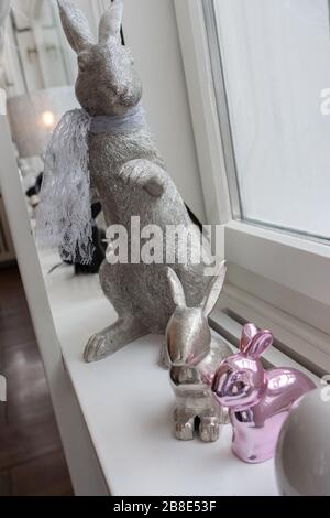 Des lapins de pâques argentés assis sur le bord latéral. Vue en grand angle des lapins assis devant une fenêtre blanche et lumineuse. Composition métallique pour le concept Holiday. Banque D'Images