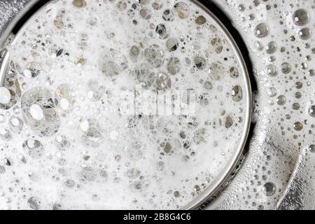 Mousse blanche avec bulles de nettoyant dans un lavabo, vue macro. Vidanger le trou avec des bulles de savon dans l'évier métallique. Bouchon de vidange réglable mécaniquement. Banque D'Images