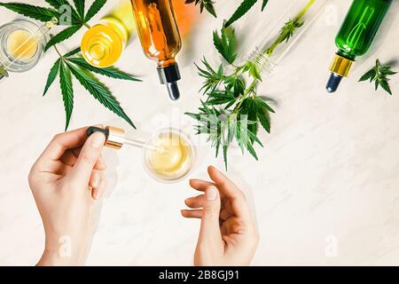 Pipeter avec de l'huile cosmétique CBD dans les mains des femmes sur un fond de table avec diverses bouteilles avec de l'huile de cannabis et des feuilles de chanvre, de la marijuana Banque D'Images