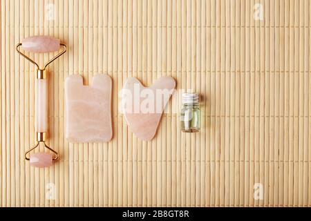 Un ensemble d'outils pour la technique de massage du visage Gua Sha fait de quartz rose naturel. Rouleau, pierre à jade et huile dans un pot en verre, sur fond de paille pour Banque D'Images
