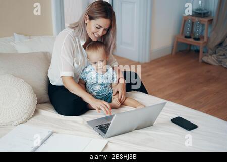 Jeune mère avec son enfant travaillant sur ordinateur portable dans la chambre à la maison. Multi-tâches, freelance et concept de la maternité Banque D'Images