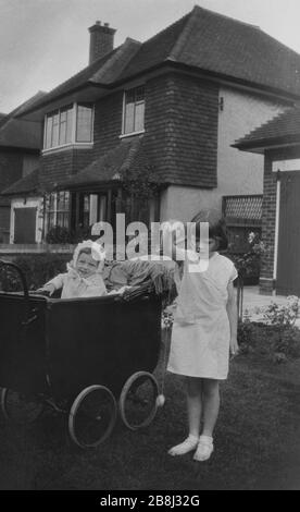 Années 1940, une jeune fille avec sa sœur, qui est assise dans un landau doublé de cuir à l'extérieur dans un jardin devant sur une rue de banlieue, Angleterre, Royaume-Uni. Banque D'Images