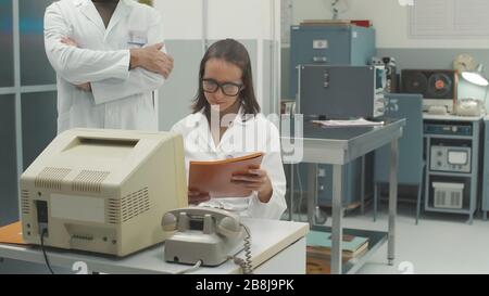 Équipe scientifique travaillant en laboratoire à l'aide de machines et d'ordinateurs anciens, le scientifique en chef vérifie le travail d'un chercheur Banque D'Images