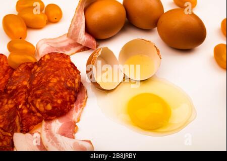 Un œuf de poulet cassé, des tranches de saucisses et de bacon tranchés et des tomates sur fond blanc. Gros plan Banque D'Images