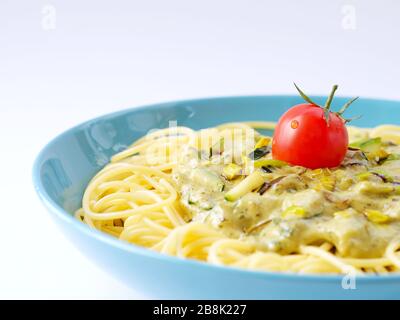 Spaghetti avec une sauce crème avec courgettes et poireaux avec quelques tomates pour la décoration dans une assiette bleu clair sur un fond blanc avec espace de blan. Banque D'Images