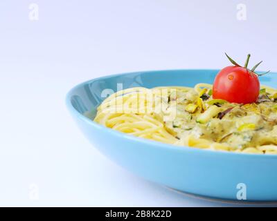 Spaghetti avec une sauce crème avec courgettes et poireaux avec quelques tomates pour la décoration dans une assiette bleu clair sur un fond blanc avec espace de blan. Banque D'Images