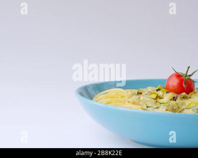 Spaghetti avec une sauce crème avec courgettes et poireaux avec quelques tomates pour la décoration dans une assiette bleu clair sur un fond blanc avec espace de blan. Banque D'Images