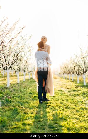 Photo de charmante jeune femme blonde dans une robe de mariage de luxe portée par son beau homme dans le jardin fleuri de printemps. Couple s'amuser Banque D'Images