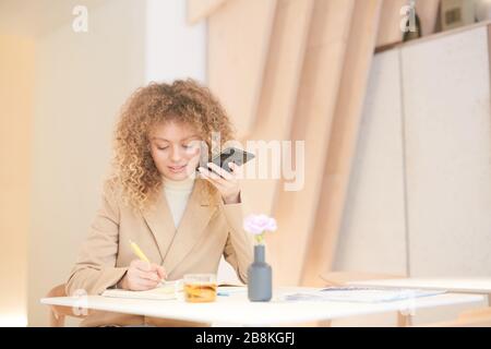 Portrait aux tons chauds de la jeune femme aux cheveux bouclés à l'aide d'un smartphone et de l'écriture pendant que vous travaillez ou que vous étudiez assis à la table dans un café, copiez l'espace Banque D'Images