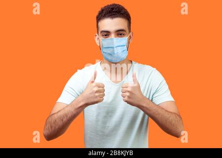 Comme ! Portrait d'un homme optimiste avec masque médical dans un t-shirt blanc montrant des pouces vers le haut et sourire, signifiant bien fait, bon travail. indoo Banque D'Images