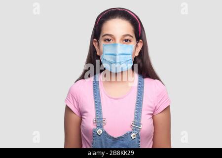 Portrait d'une fille avec masque médical dans un style décontracté, t-shirt rose et salopettes en denim bleu debout et regardant. Plan de studio intérieur, isolé sur ba gris Banque D'Images