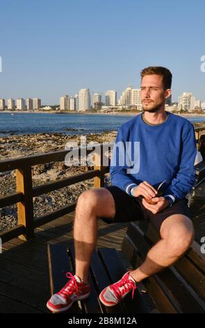 Un bel homme buvant Yerba Mate Infusion dans la rambla de Punta del Este, Maldonado, Uruguay avec l'océan atlantique et la ville à l'arrière-plan Banque D'Images