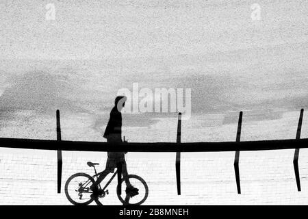 Silhouette d'ombre d'un jeune homme marchant et poussant son vélo sur la rue vide de la ville, en noir et blanc Banque D'Images