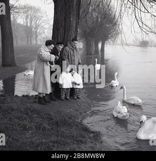 1964, historique, famille avec enfants et grand-mère dans leurs manteaux extérieurs se nourrissant des canards par un lac débordant, Angleterre, Royaume-Uni. Les dames portent des manteaux de fourrure, tandis que le mari porte un manteau de sport, avec les deux jeunes filles en costume assorti. Banque D'Images