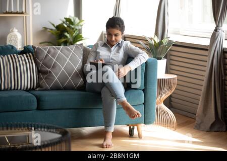 Jeune femme indienne utilisant une tablette informatique, assise sur un canapé confortable Banque D'Images