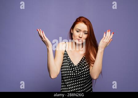 Portrait d'une jeune femme caucasienne magnifique avec des cheveux branchés et des émotions confuses, se propageant les mains. Je ne comprends pas et je ne sais pas. Fille dans une robe noire à pois sur un fond mauve. Banque D'Images